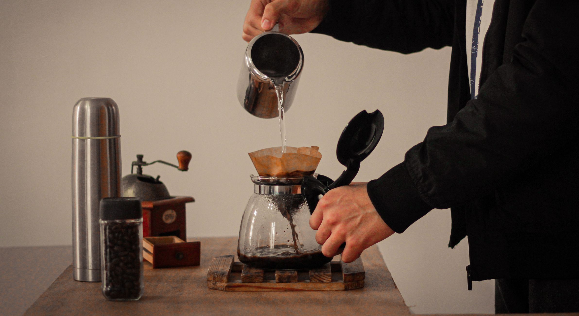 A barista pours hot water through a coffee filter into a glass pot, surrounded by coffee-making tools.