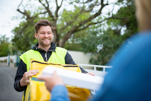 Happy delivery man in yellow vest hands over a package outdoors, smiling cheerfully.