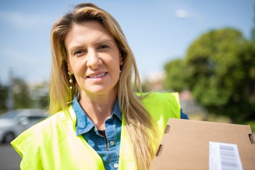 Happy delivery woman holding a package outdoors in a sunny environment.