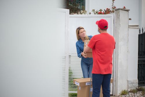 Free A delivery man hands a package to a woman at a metal gate, following COVID-19 precautions. Stock Photo