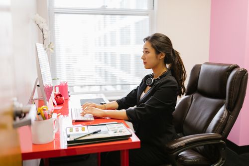 Free Professional Asian woman engaging in remote work at a stylish office desk with a computer and headphones. Stock Photo