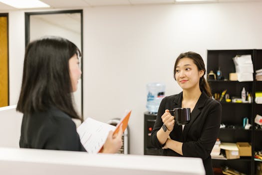 Two businesswomen conversing in an office setting, holding a coffee mug.