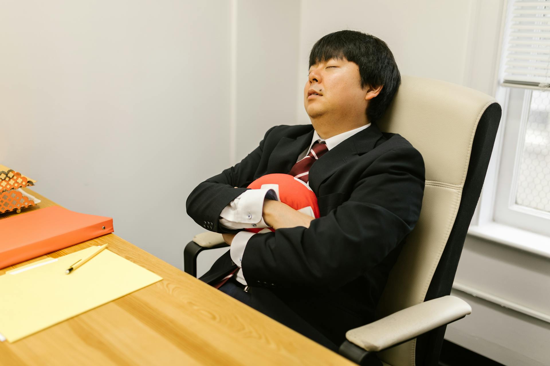 Asian businessman sleeping in office chair, arms crossed with a small pillow.