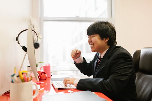 A joyful man in a suit celebrating success at his modern office desk, smiling with excitement.