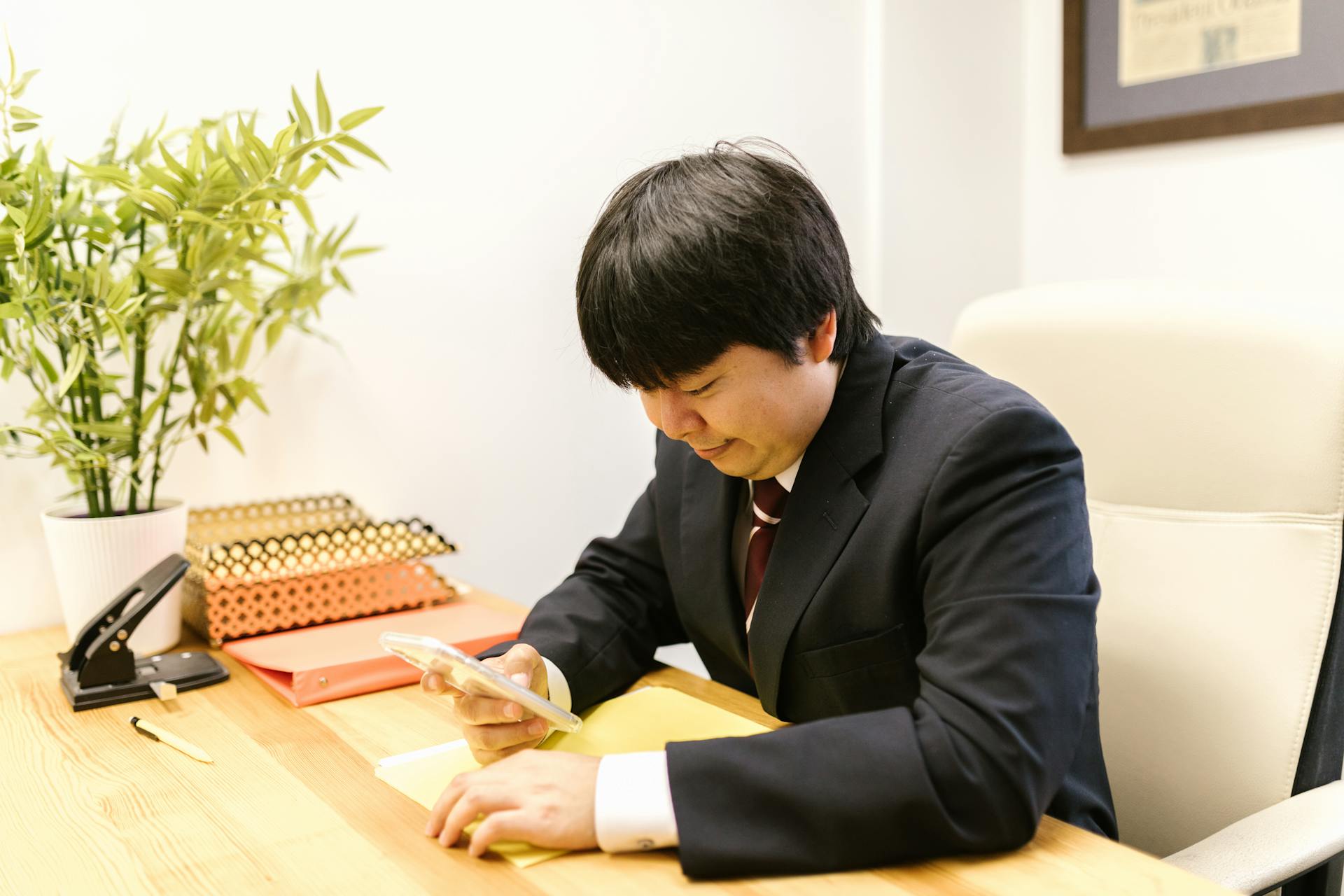 Asian man in corporate attire using a smartphone at his desk in an office setting.