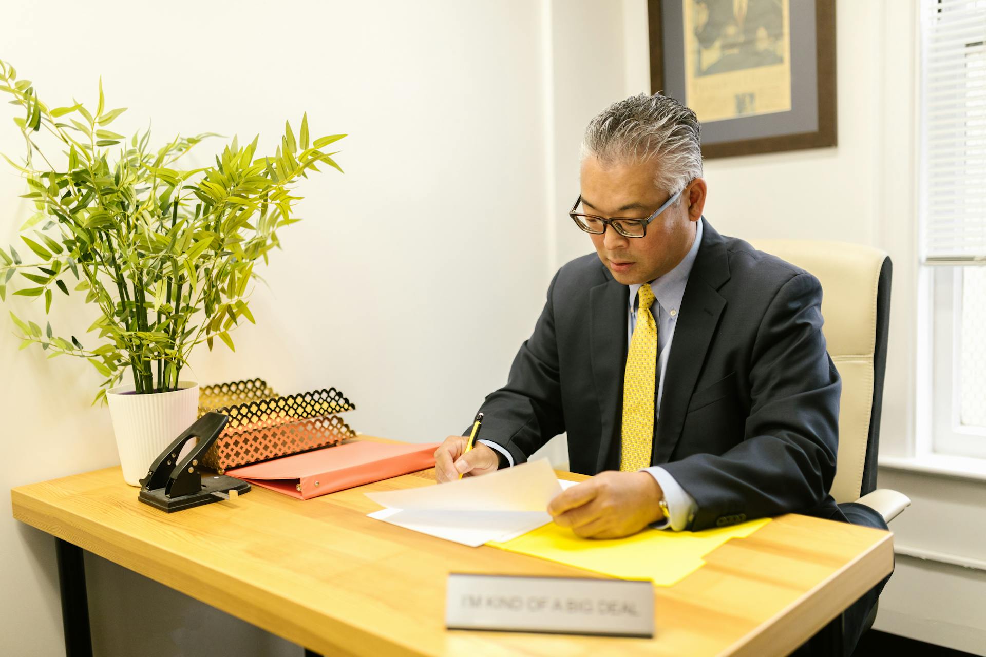 An adult man in business attire working at a desk with documents in an office environment.