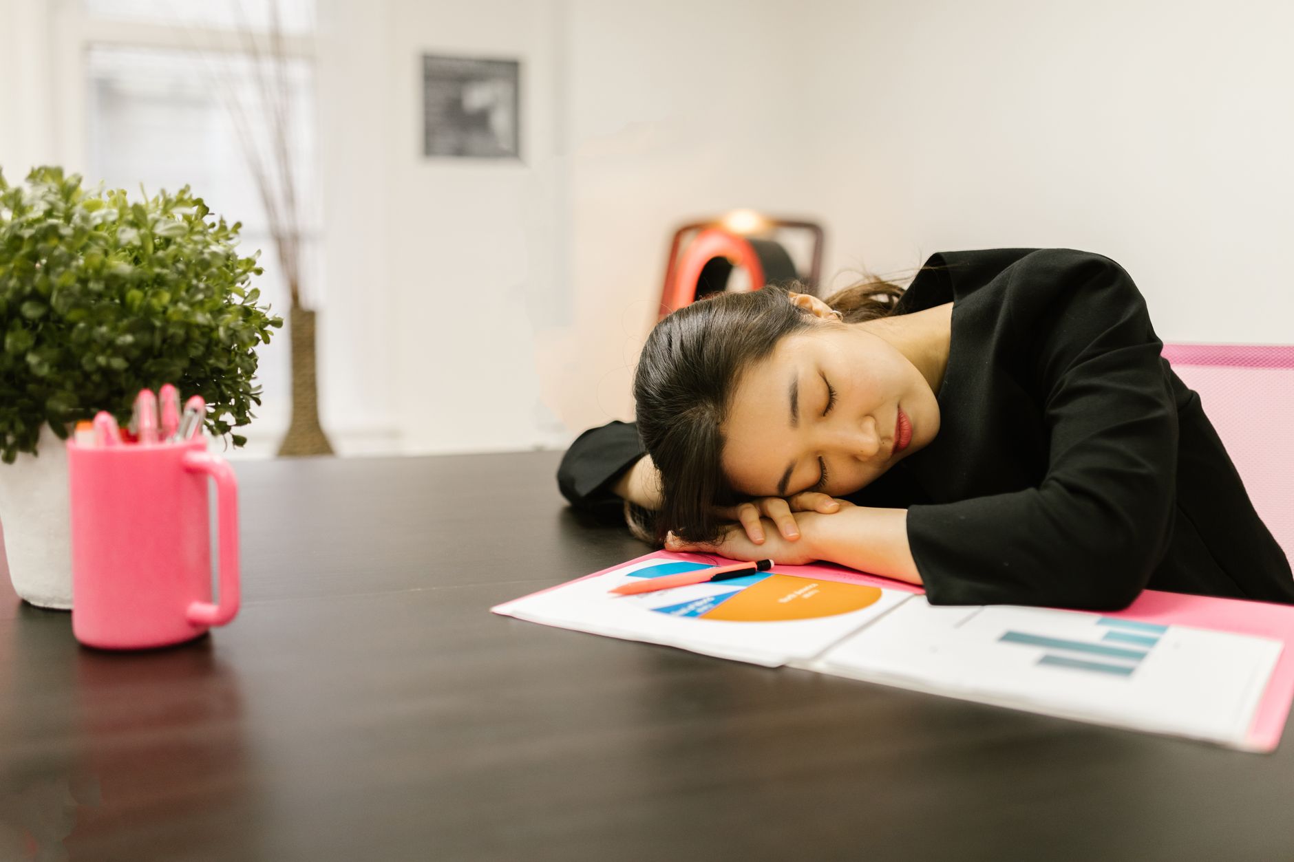 An exhausted woman in a black outfit rests her head on a desk in an office setting, surrounded by papers and a potted plant.