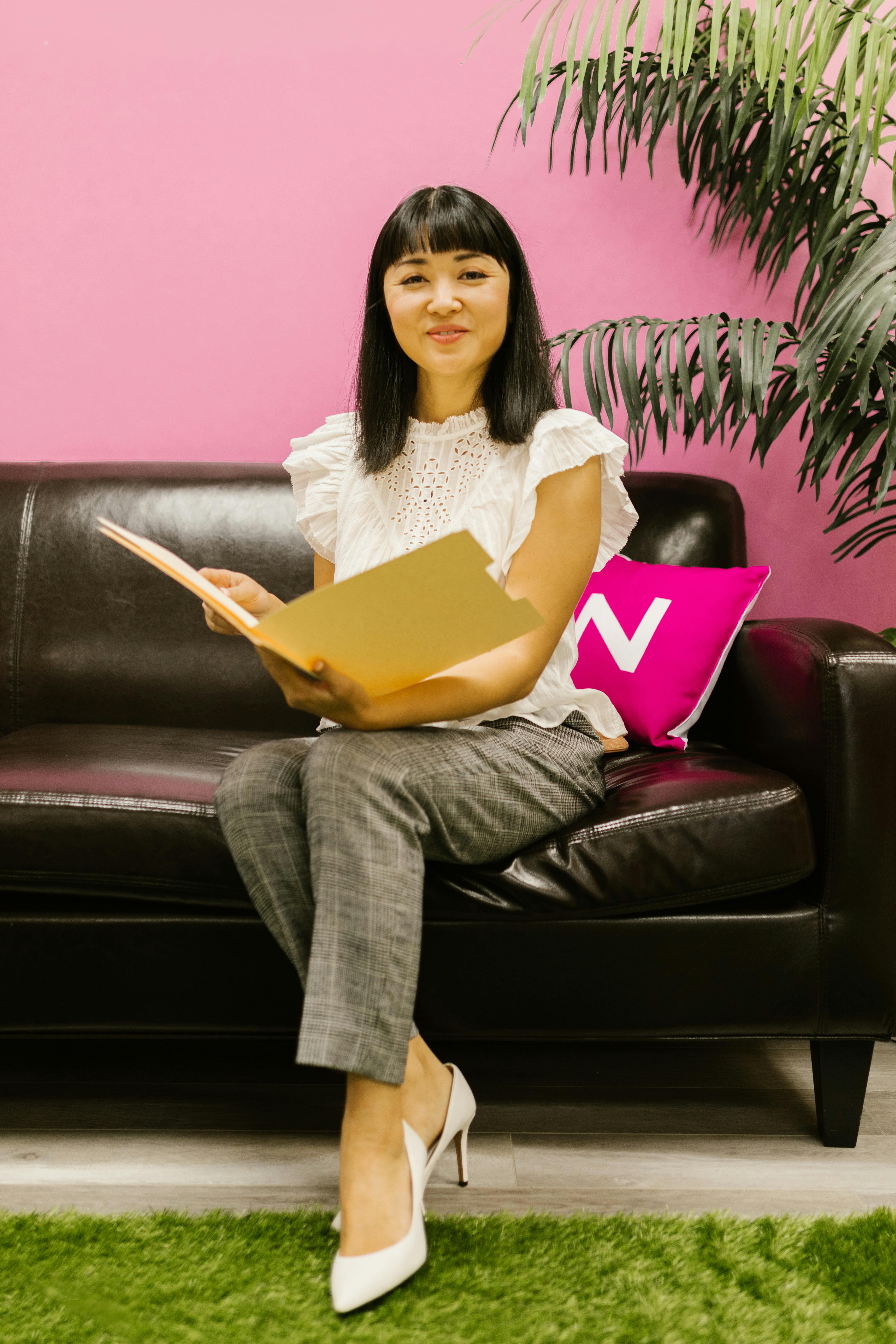 Asian woman in office attire sitting on black sofa, holding documents, and smiling in a modern office setting.