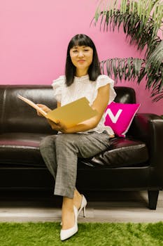 Asian woman in office attire sitting on black sofa, holding documents, and smiling in a modern office setting.