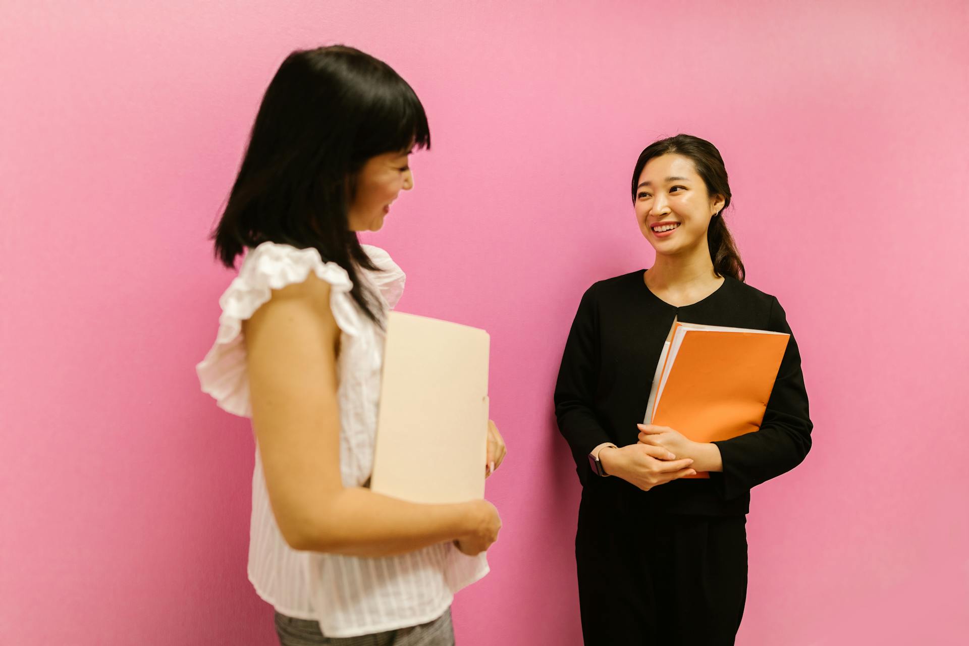 Two Asian women in business attire discussing work while holding folders against a pink background.