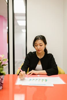 Asian businesswoman in office reviewing documents at a red desk, focused and professional.