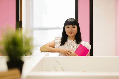 Free Asian woman in white top holding a pink notebook, smiling in a modern office setting. Stock Photo