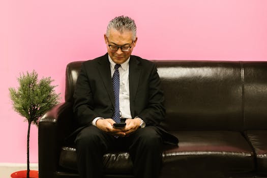 Asian man in a suit using smartphone, sitting on a leather sofa against a pink background.