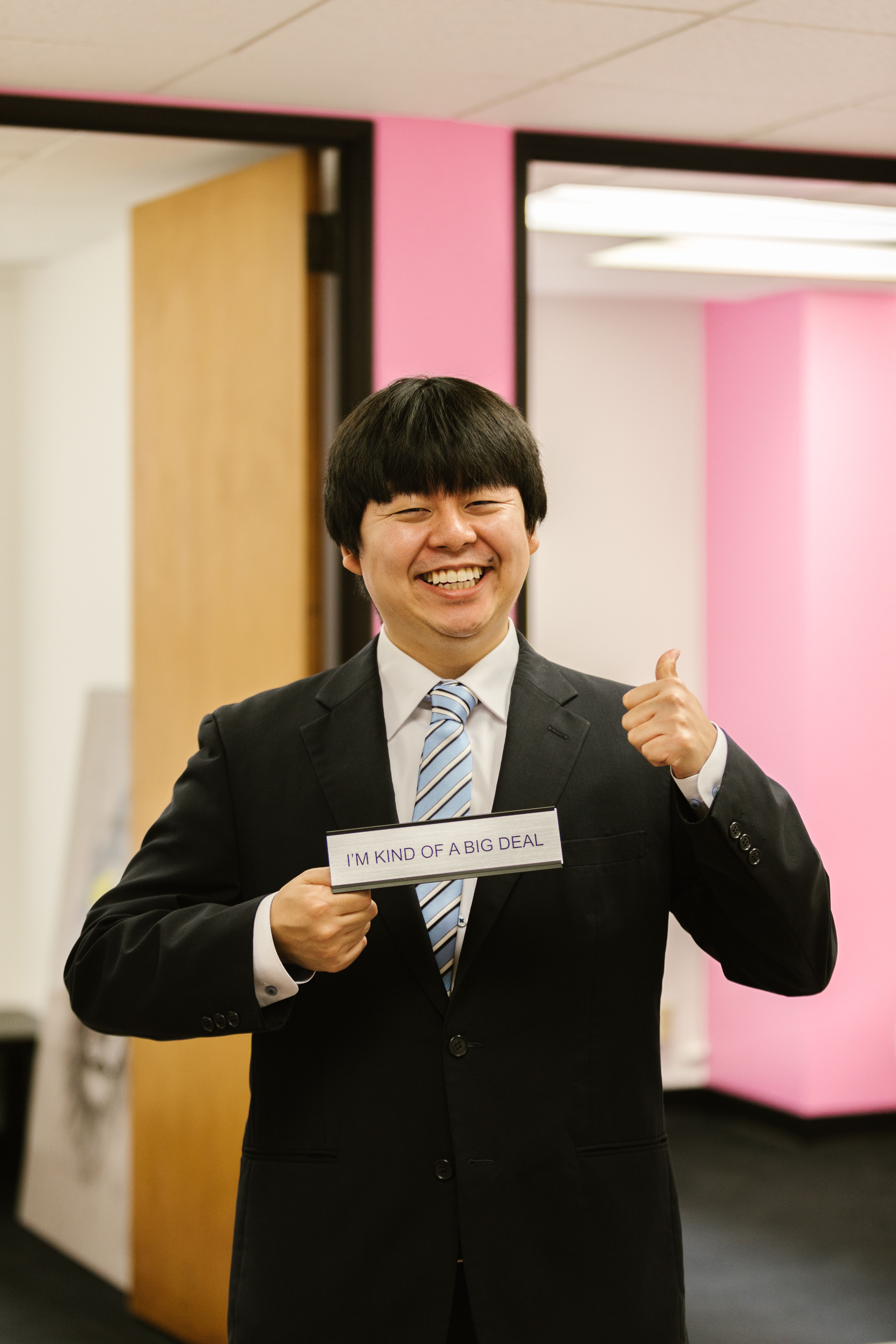 Asian businessman smiling with a humorous sign in an office setting, demonstrating confidence.