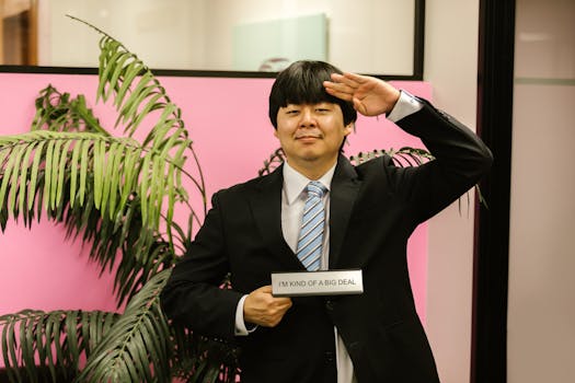 Asian man saluting in formal attire, holding a humorous sign indoors.