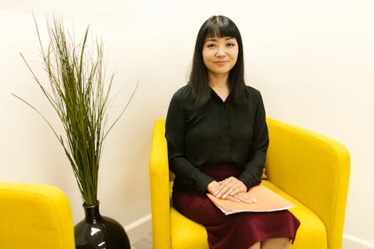 Asian woman sitting on yellow sofa with a folder in a modern office setting.