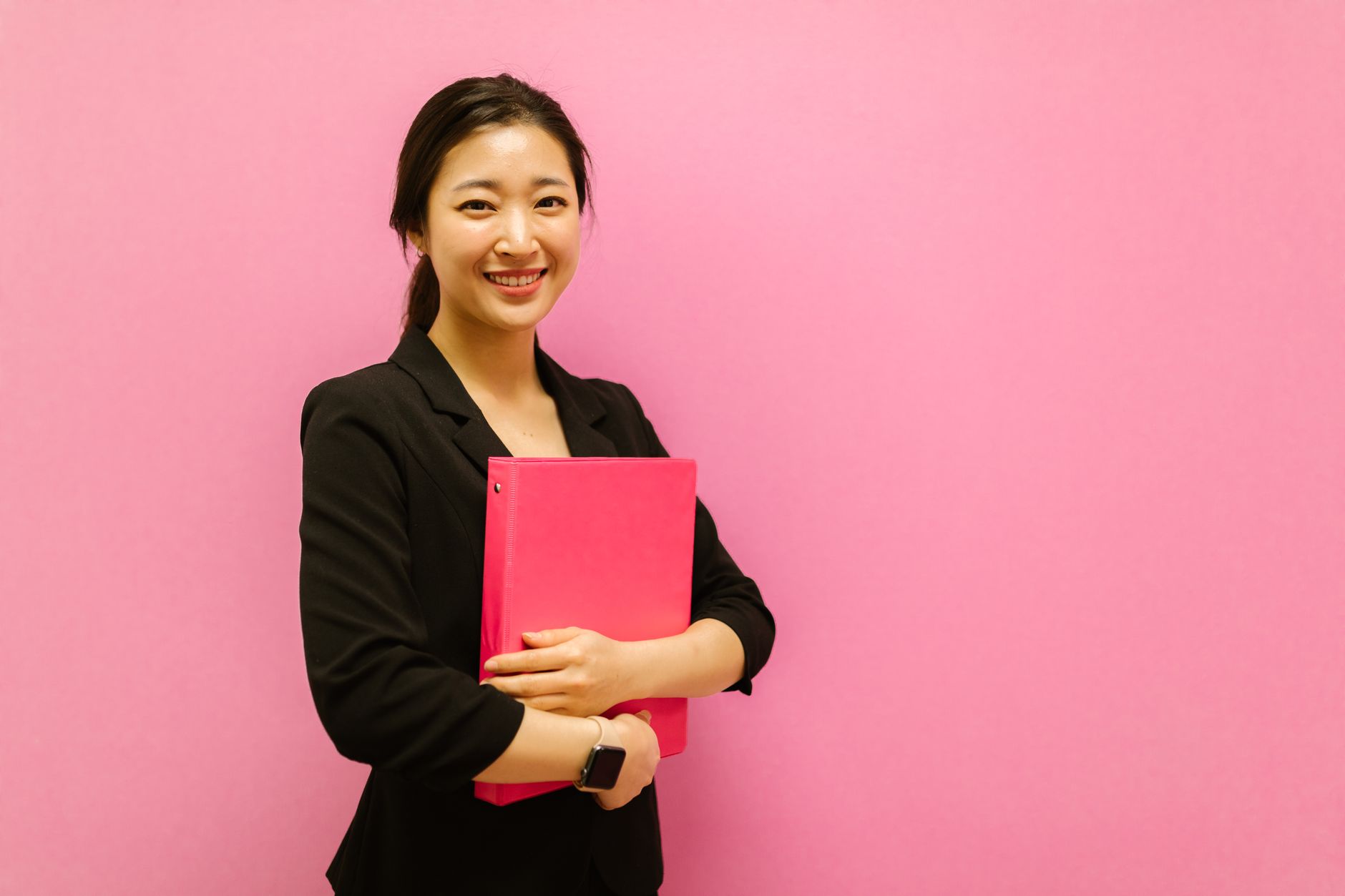 Smiling Asian woman holding a pink binder against a pink background, conveying positivity.