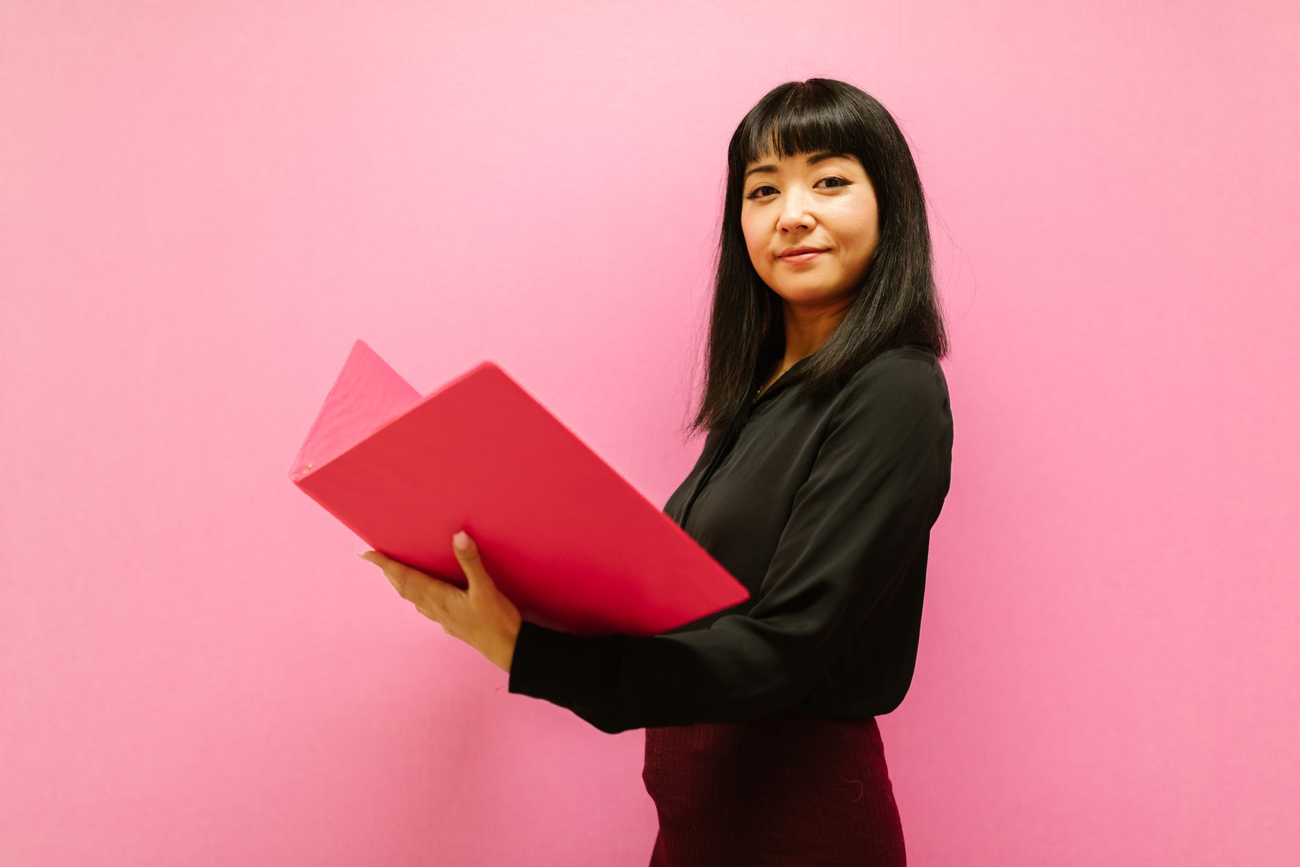 Smiling woman in business attire holding a pink folder against a pink background.