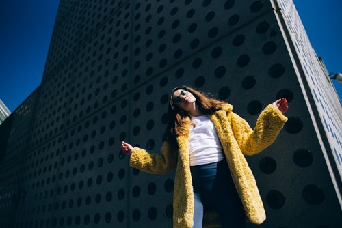 Free Stylish woman posing in a vibrant yellow faux fur coat and sunglasses against a modern urban backdrop. Stock Photo
