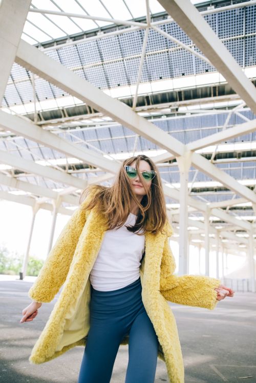 Free Trendy woman in yellow coat with sunglasses posing outdoors under a modern structure. Stock Photo