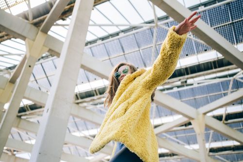 Free Fashionable woman in yellow faux fur coat and sunglasses poses under modern architecture. Stock Photo