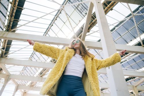 Free Fashionable woman in a yellow faux fur coat posing confidently under a modern structure. Stock Photo