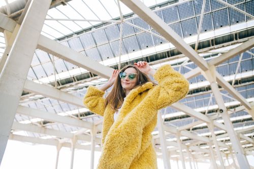 Free A fashionable woman poses confidently in a yellow fuzzy coat and sunglasses under a modern structure. Stock Photo