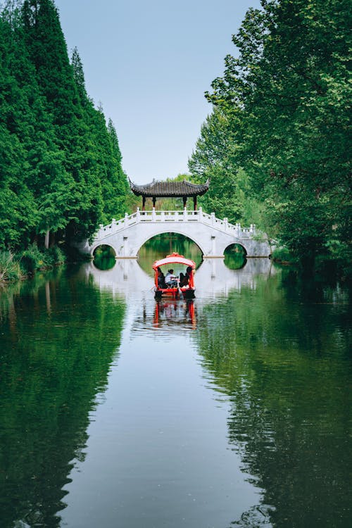 Free Oriental pleasure boat with tourists floating on peaceful water of lake near footbridge in greenery terrain Stock Photo