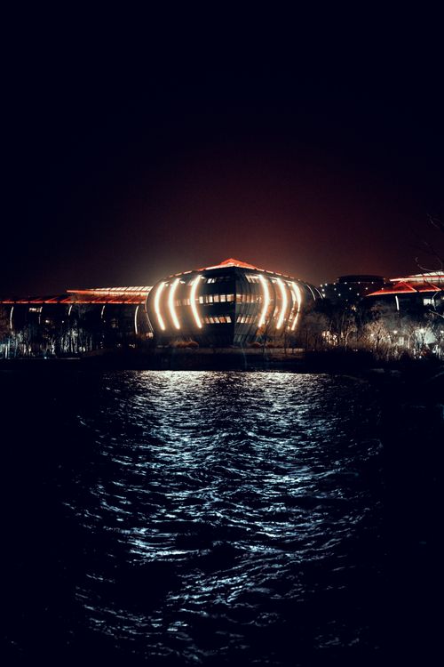 Free A modern building glowing with lights, reflected in dark water under a night sky. Stock Photo