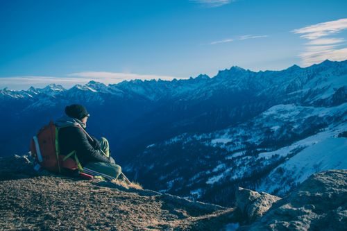 Free A man sits on a mountain ridge enjoying a breathtaking snow-capped mountain view under a clear blue sky. Stock Photo