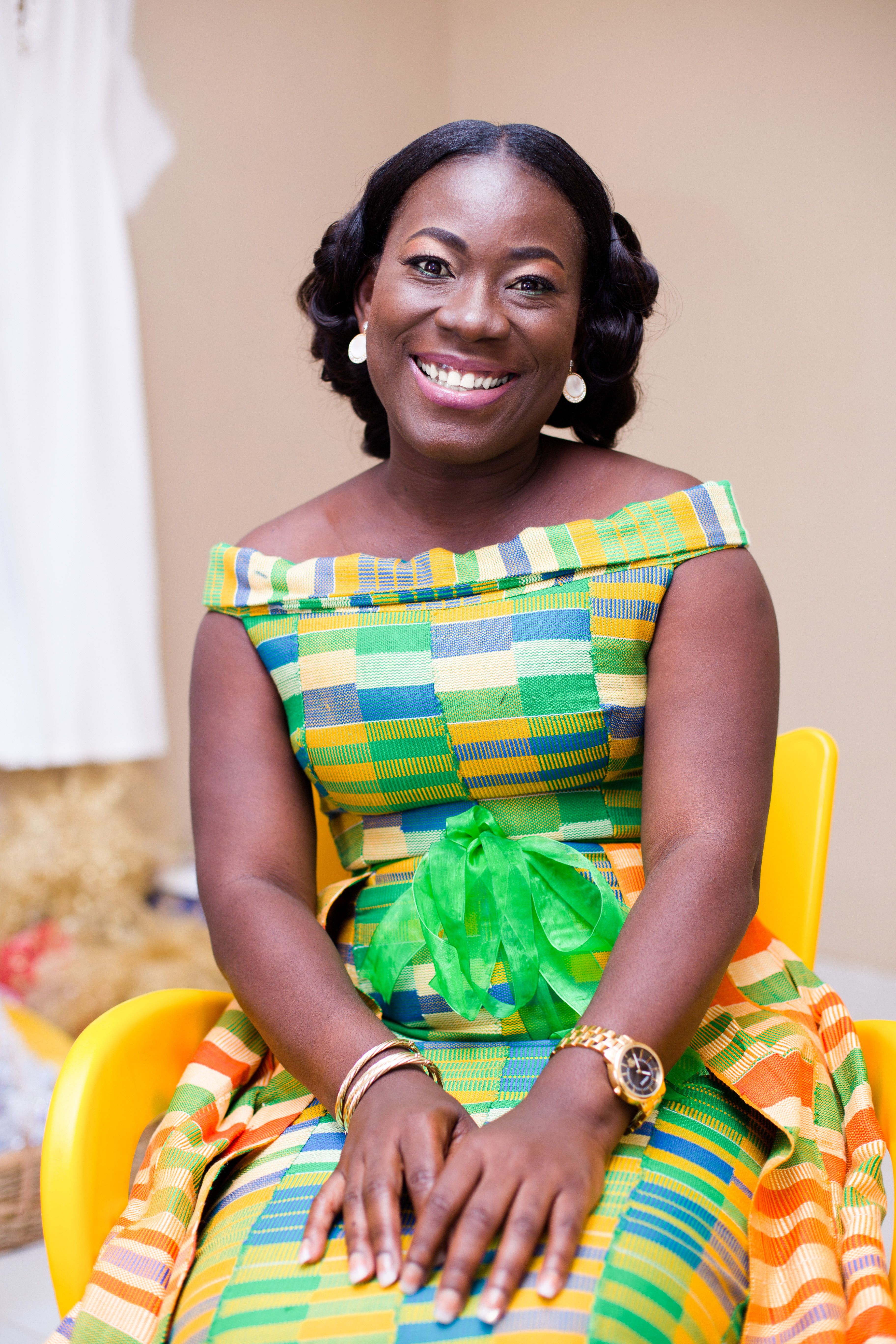 Black woman smiling, wearing vibrant traditional dress indoors.