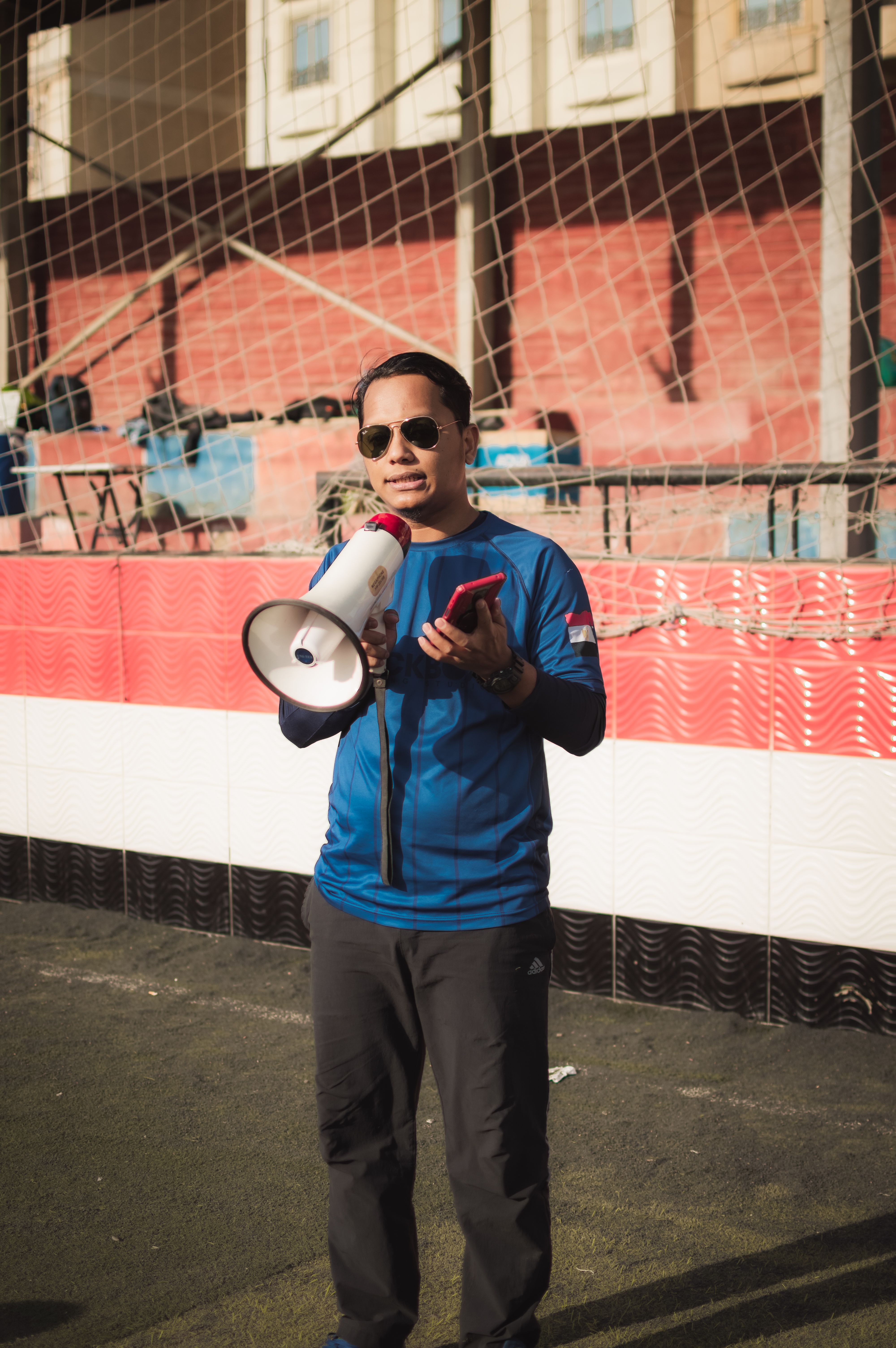 Person in sunglasses holding megaphone and smartphone on outdoor sports field.