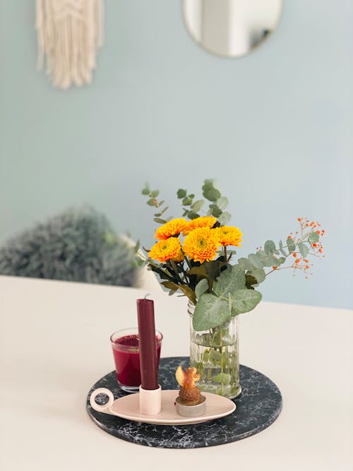 Composition of bunch of bright yellow marigolds flowers and green twigs and leaves aromatic candles of maroon color placed on tray on white table in light room