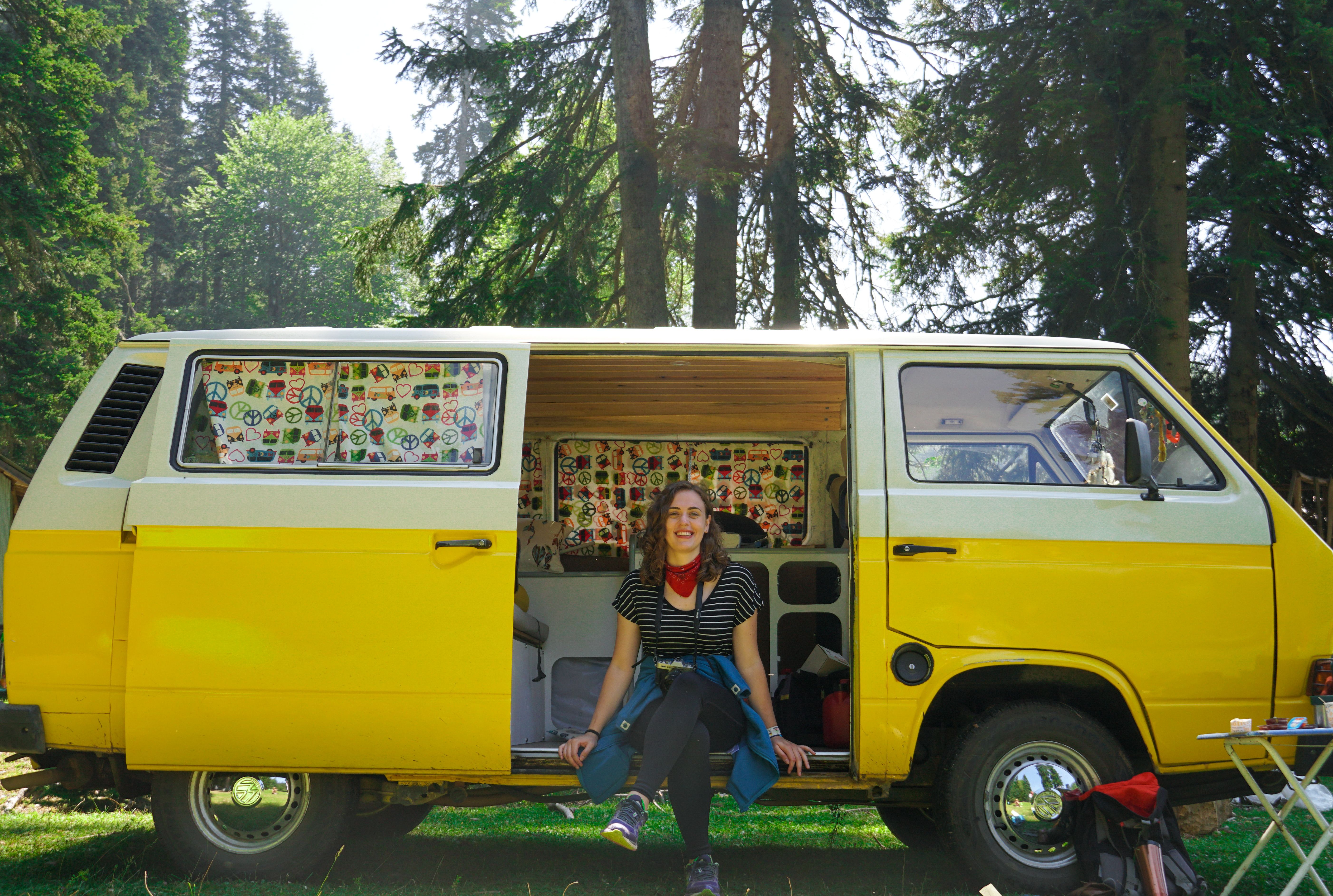 Happy woman sitting in yellow camper van amidst lush forest, embracing outdoor adventure.