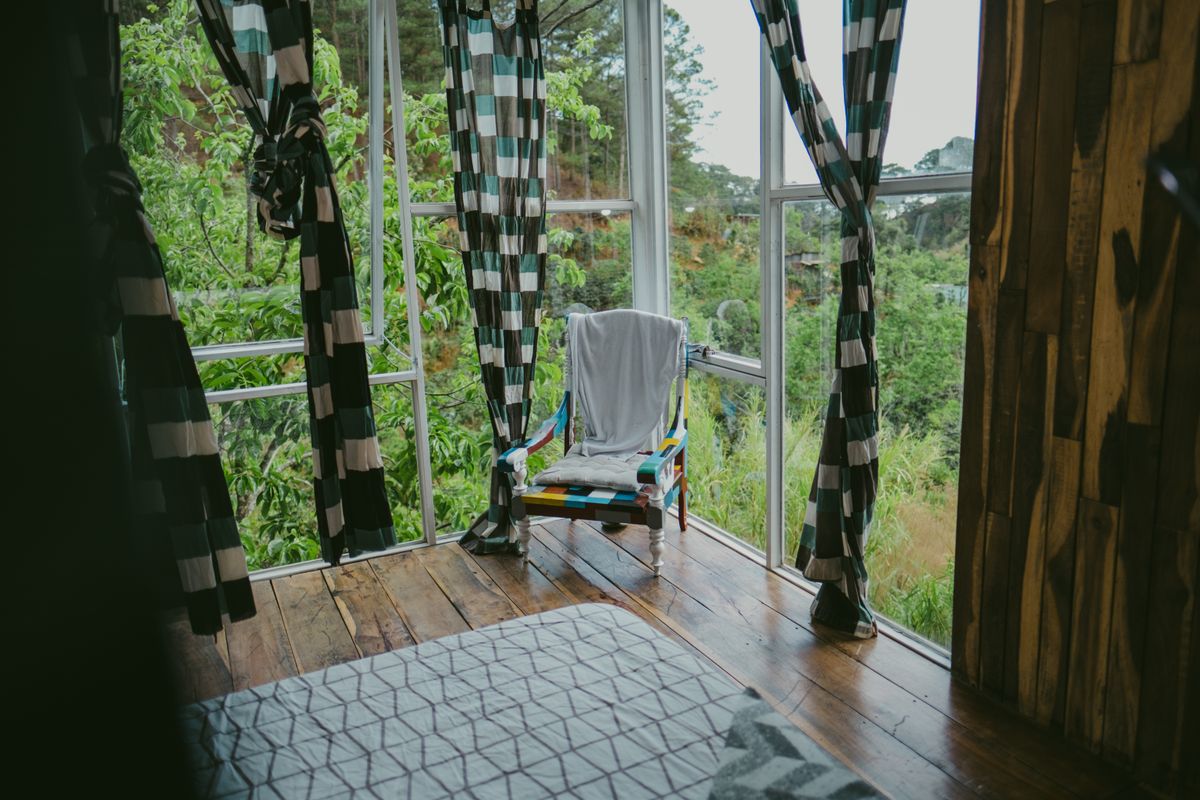 A cozy room with a colorful chair, checkered curtains, and a lush green view through large windows. Stock Photo.