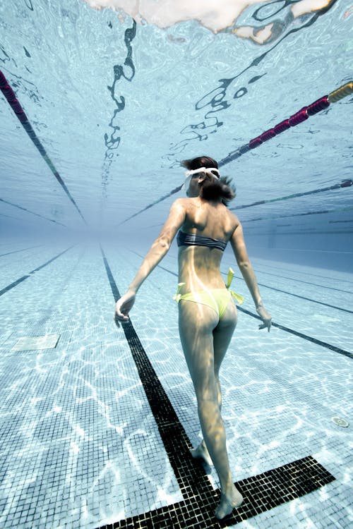 Free Back view of anonymous female in swimsuit and diving mask swimming underwater in clear water of swimming pool Stock Photo