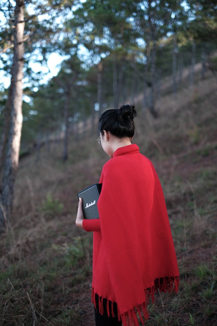 A Person In A Red Shawl Holding A Bluetooth Speaker In The Woods