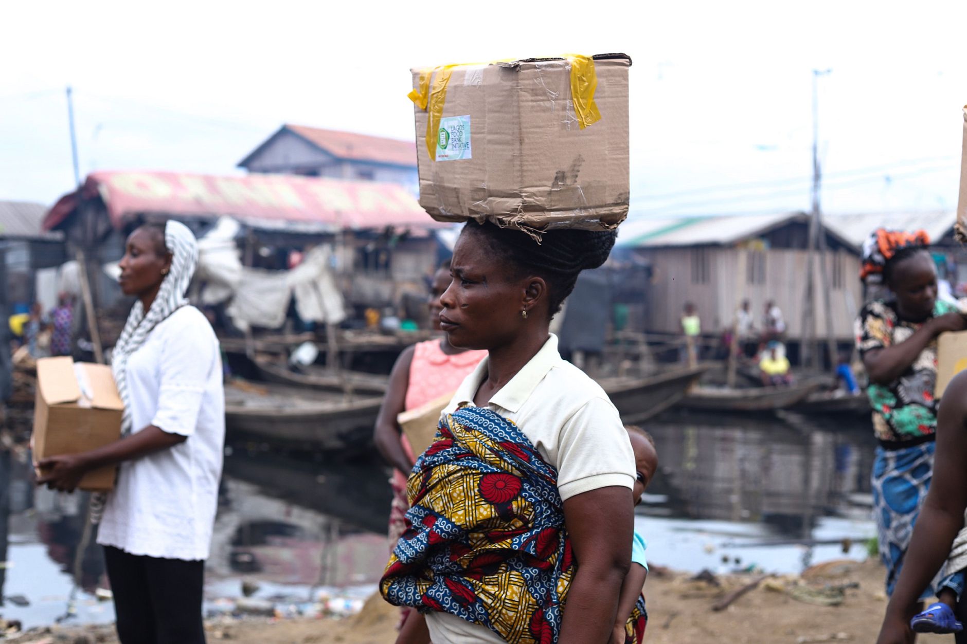 African women in traditional attire carrying boxes by a riverside market area.