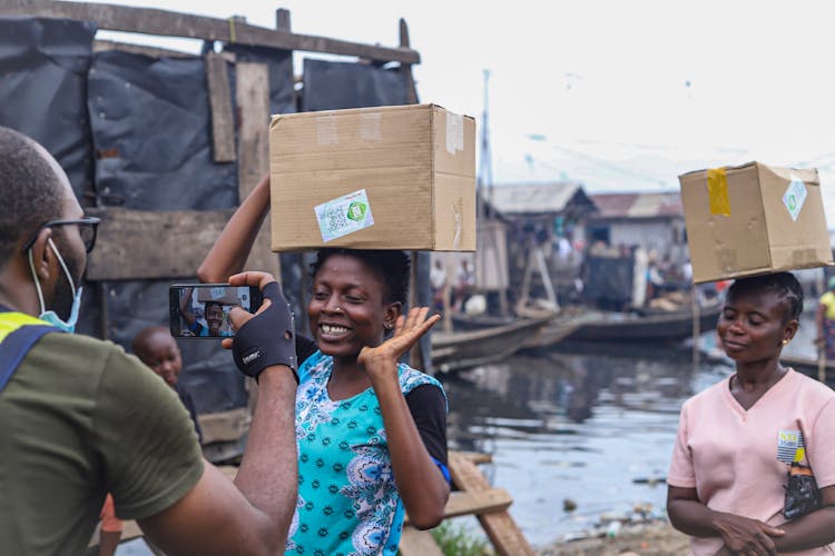 Man Photographing Smiling Women Carrying Cardboard Boxes On Top Of Their Heads 