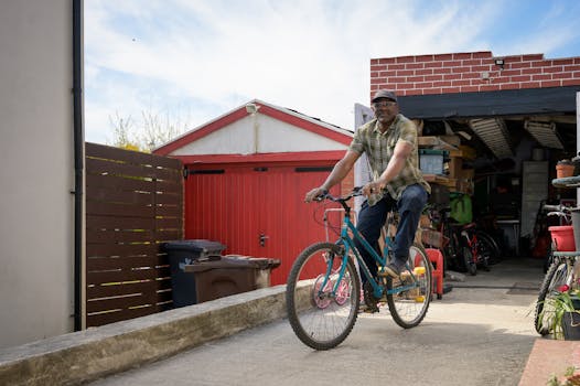 Smiling man rides a bike near an open garage on a sunny day.