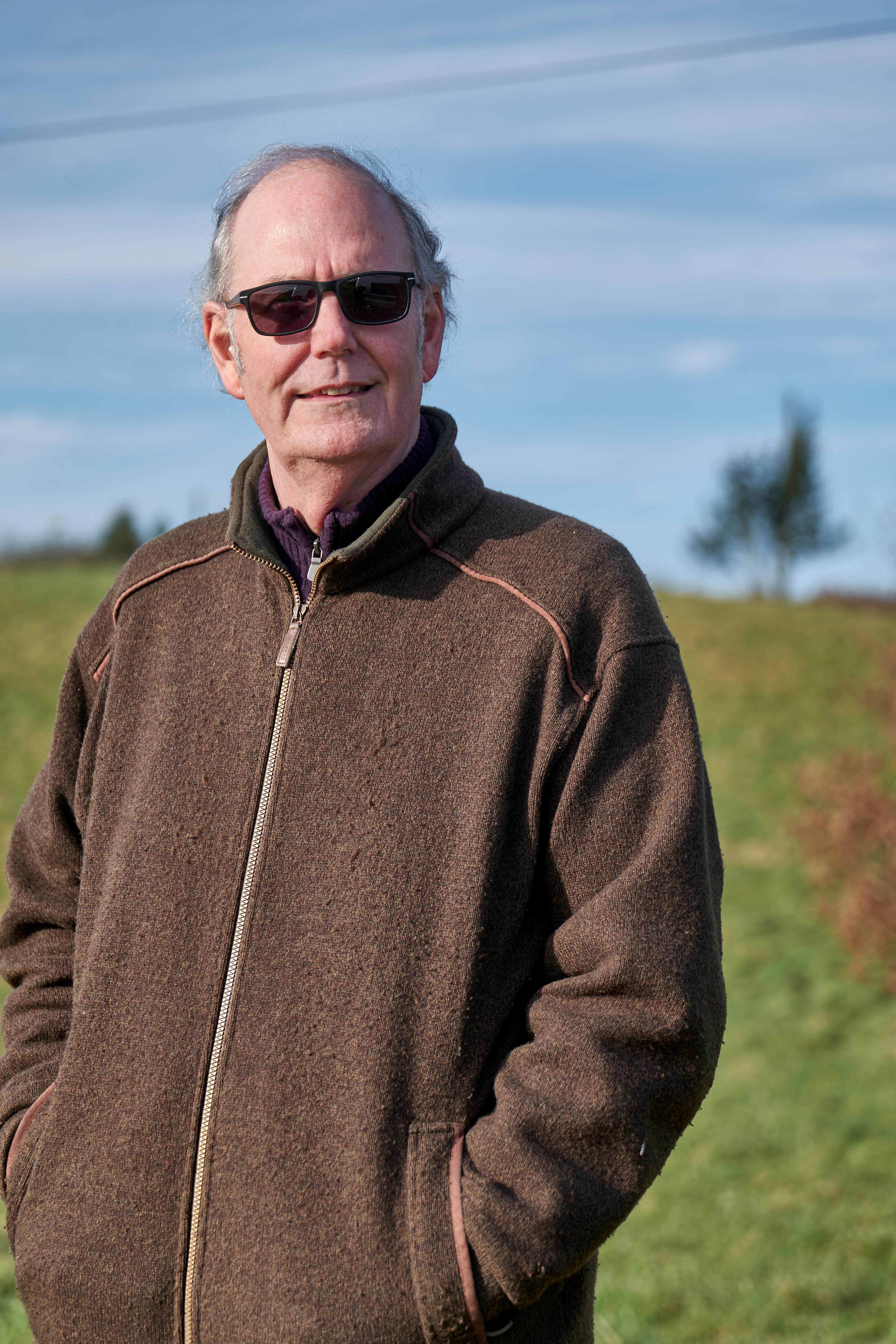 Senior man with sunglasses stands confidently outdoors on a sunny day, hands in pockets.