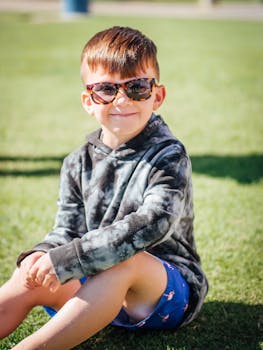 A cheerful young boy wearing sunglasses sits on the grass, enjoying a sunny day outdoors.
