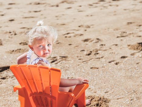 Adorable blond child sitting on an orange chair at the sandy beach enjoying a sunny day.