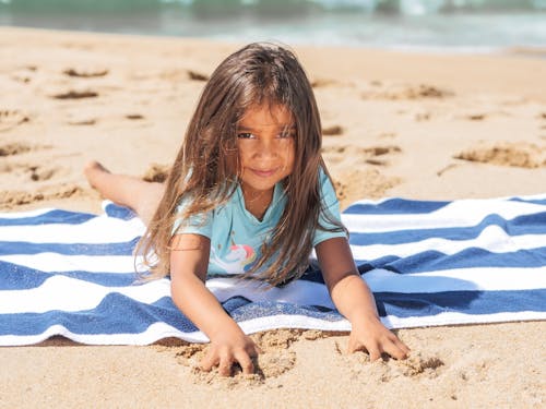 Girl in Blue Shirt Sitting on White and Blue Stripe Textile on Beach