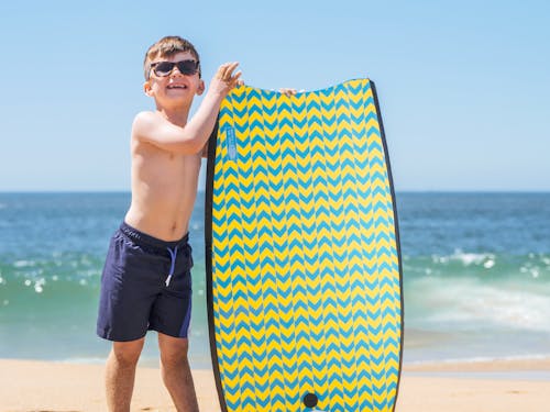 grátis Homem De Short Preto Segurando Uma Prancha De Surfe Azul E Branca Na Praia Foto profissional
