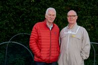 Man in Red Zip Up Jacket Standing Beside Man in Brown Button Up Shirt