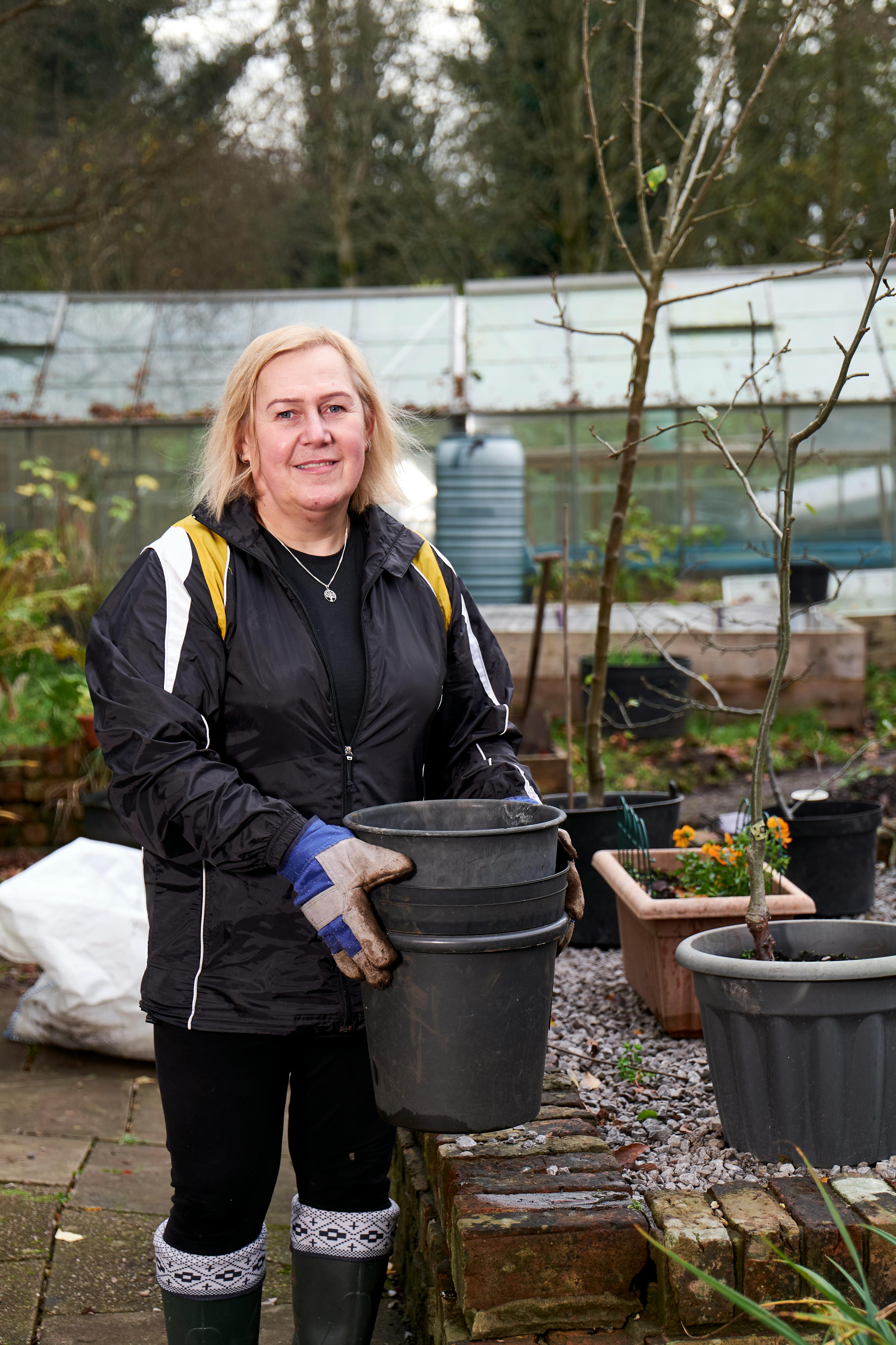 Woman gardener in a garden holding pots. Greenhouse and plants in the background.