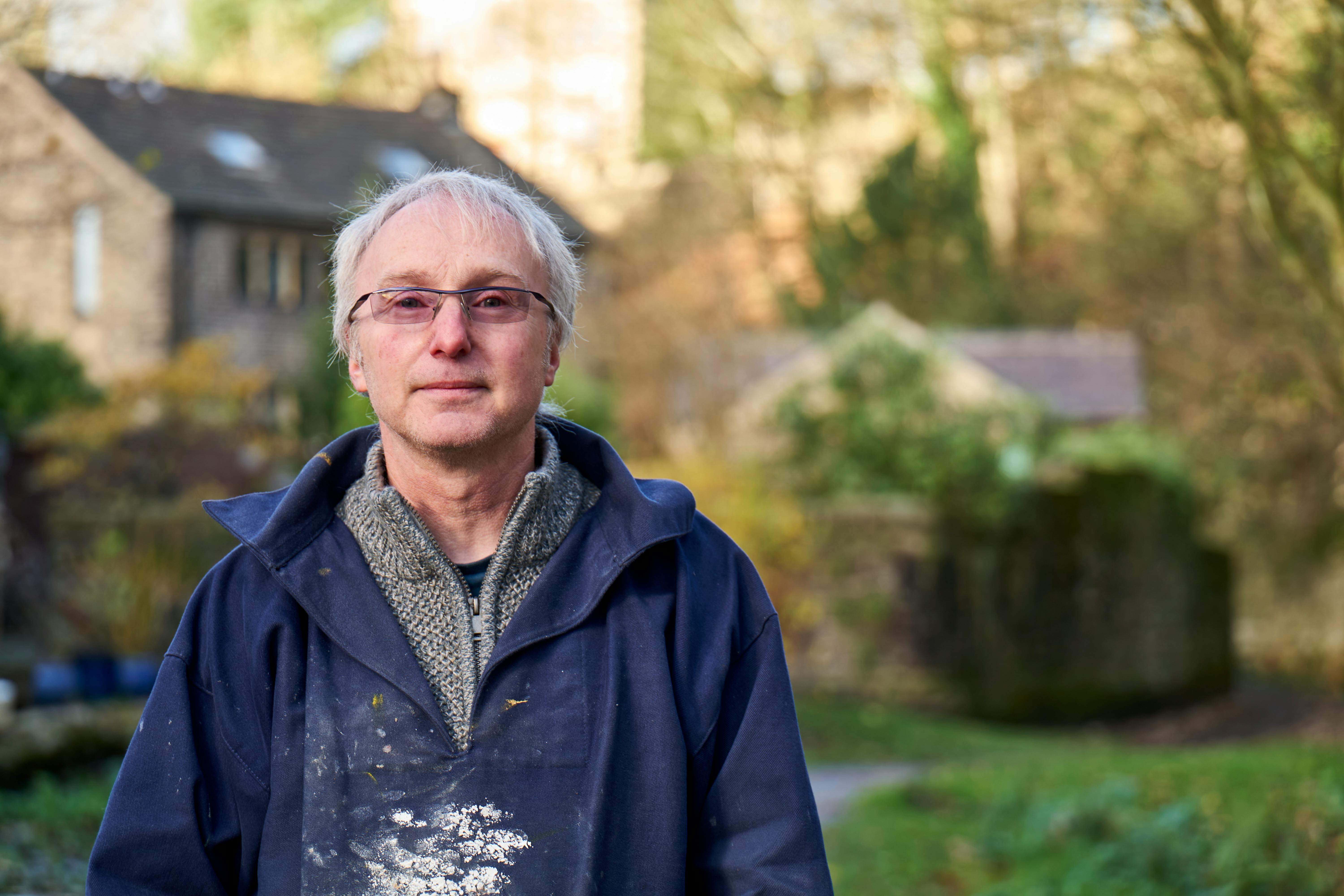 Portrait of a senior man wearing glasses and a blue jacket standing outdoors with a house in the background.