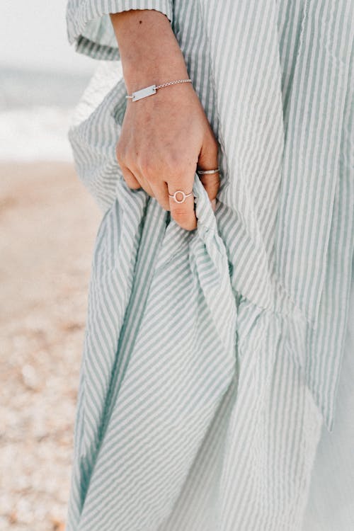 Female hand with ring and bracelet holding cotton light dress on background of sandy seashore in sunny day