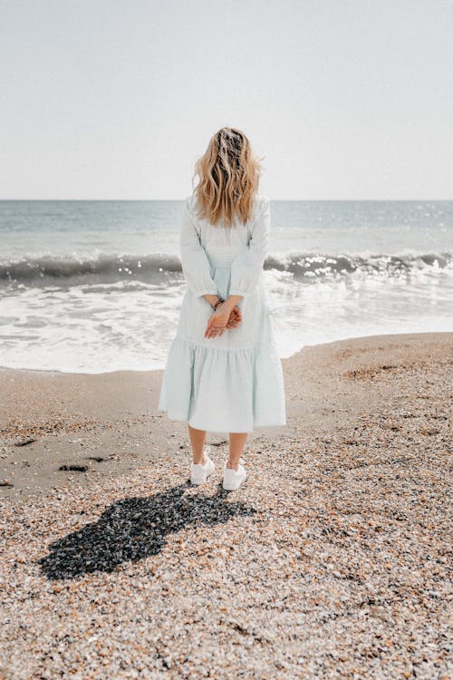 Full body back view of female wearing light blue dress and white sneakers standing with crossed arms on sandy coastline and looking at wavy foam of ocean in sunlight
